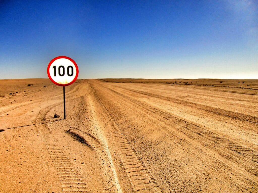 Camino abierto en el desierto con una señal de límite de velocidad que muestra 100 bajo el cielo despejado.