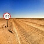 Camino abierto en el desierto con una señal de límite de velocidad que muestra 100 bajo el cielo despejado.