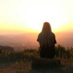 Mujer sentada en la naturaleza, viendo la puesta de sol sobre los campos. Escena al aire libre pacífica y serena.
