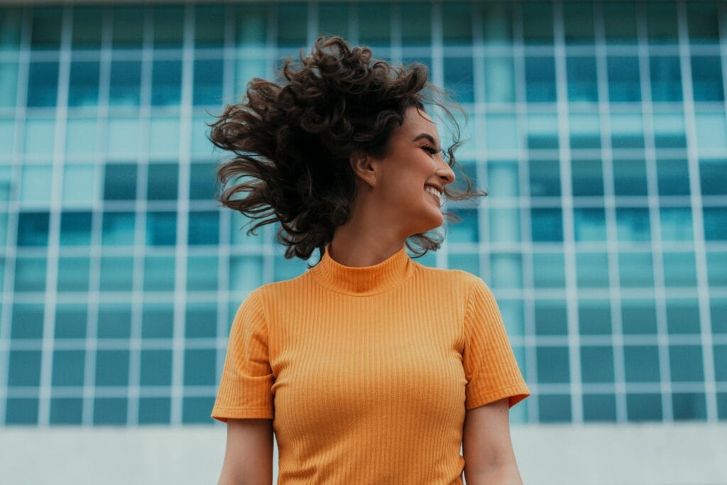 Mujer alegre sonriendo y volteándose el pelo frente a un edificio moderno.