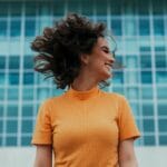 Mujer alegre sonriendo y volteándose el pelo frente a un edificio moderno.