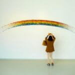 Una mujer está de pie observando una colorida instalación de arte del arco iris en el interior.